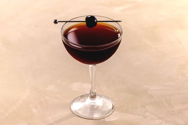 Black Manhattan cocktail in a cup with an brown and red color twist and a cherry